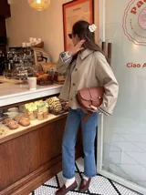 Chaqueta corta de gabardina en color beige, con cuello camisero, cierre frontal de botones y mangas largas con puños vueltos que revelan un forro a cuadros.