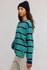 Remera manga larga de algodón con diseño de rayas horizontales en tonos verdes, azul marino, rosado y celeste. Presenta cuello redondo y un pequeño parche decorativo en el borde inferior.