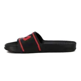 Chancletas tipo slide negras con tira ancha que presenta el logo Fila en rojo, bordeado por una línea roja. La plantilla es texturada.