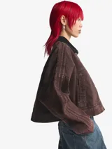 Chaqueta de lona de algodón en color marrón con efecto desgastado, corte holgado, cuello camisero y cierre frontal con botones metálicos. Presenta un pequeño logo de Adidas en tono dorado en el pecho y detalles de costuras decorativas en las mangas.