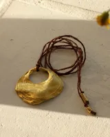 Collar artesanal con dije central de bronce macizo con textura orgánica irregular, suspendido de un cordón largo de hilo de seda natural. El diseño permite diversas formas de uso, incluyendo atado al cuello o como gargantilla.