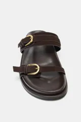 Sandalia plana de mujer color marrón oscuro, con dos tiras anchas de ante ajustables mediante hebillas metálicas doradas. Presenta una suela gruesa y plana.