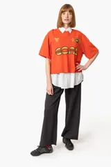 Remera naranja con cuello polo blanco, estampado frontal con la palabra "GOAL" en amarillo y negro, y logos bordados.