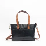 Cartera tipo tote de cuero sintético negro con detalles en color suela, incluyendo asas de mano y correa larga ajustable. Presenta un bolsillo frontal externo y herrajes metálicos.