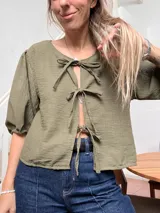 Blusa de lino color verde militar, con mangas cortas abullonadas y cierre frontal mediante tres lazos decorativos tipo moño.