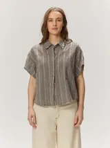 Camisa holgada de manga corta y cuello camisero, confeccionada en mezcla de lino y algodón con diseño de rayas verticales en tonos grises y marrones. Presenta detalles de costuras decorativas en rojo y botones variados en el cuello.