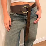 Cinto ancho de cuero marrón con diseño asimétrico, que presenta un aro metálico dorado como elemento central y una tira de gamuza larga que se anuda para el cierre.