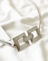 Cinturón plateado con hebilla geométrica formada por dos cuadrados unidos por una barra horizontal. El cinturón es elástico y ajustable.