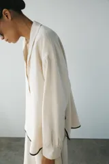Camisa de lino color crudo con cuello solapa, manga larga y bajo asimétrico. Presenta detalles de ribetes negros a contraste y aberturas laterales con tiras para ajustar.