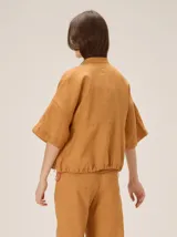 Chaqueta de lino y rayón color tostado, de corte holgado, mangas cortas amplias, cuello mao y cierre frontal oculto con cremallera. Presenta pespuntes a contratono en las costuras y cordón ajustable en el bajo.