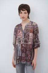 Camisa de corte recto con cuello mao y cierre frontal parcial de botones. Presenta un diseño estampado tipo patchwork con motivos florales y geométricos en tonos tierra, marrones y beige. Posee mangas tres cuartos con puños simples.
