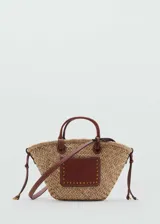 Bolso tipo canasta de paja con aplique de cuero marrón con tachas doradas, doble asa de mano y correa bandolera desmontable.