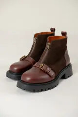 Bota de caña corta combinada en cuero liso y gamuza color marrón. Presenta cierre frontal con cremallera, tira con hebilla metálica decorativa en el empeine y suela track de goma negra.