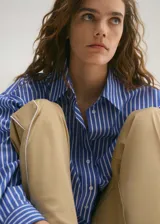 Camisa celeste a rayas verticales blancas, de corte oversize y largo crop. Tiene cuello camisero, cierre frontal con botones, manga larga con puños abotonados y un bolsillo de parche en el pecho.