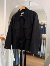 Campera negra de algodón de silueta relajada, con cuello amplio, bolsillos delanteros con tapa y cinturón ajustable del mismo género.