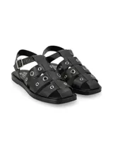 Sandalia tipo cangrejera de cuero graneado color negro, con diseño de tiras cruzadas decoradas con ojales metálicos. Cuenta con tira ajustable al tobillo con hebilla metálica y suela plana de PVC.