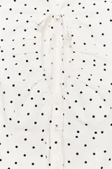 Top de popelina de color blanco con estampado de lunares negros, cuello mao con escote en pico y lazada, mangas cortas abullonadas y cierre frontal con botones.