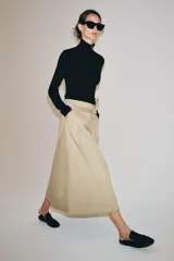 Falda midi color beige confeccionada en hilatura con mezcla de algodón. Presenta tiro medio, cintura con trabillas, bolsillos laterales ocultos en costura y cinturón del mismo tejido. Su diseño incluye un bajo acabado en línea evasé y cierre lateral con cremallera.