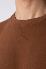 Remera de corte oversized en color marrón, confeccionada en tejido de algodón con textura de micropanal. Presenta cuello redondo con detalle de costura en forma de X y mangas cortas de estilo caído.
