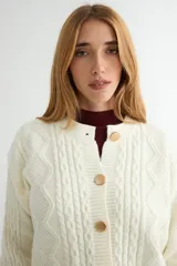 Cardigan de punto trenzado color crema con cuello redondo y cierre frontal de botones grandes en tono dorado. Presenta mangas largas con puños acanalados y un diseño de tejido con textura.