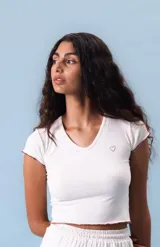 Top corto de manga corta, color blanco jaspeado, con escote en V y ribete de costura en color rojizo/marrón en el cuello y los bordes de las mangas. Presenta un pequeño bordado de corazón en el pecho.