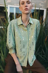 Camisa de corte recto en color verde claro con estampado de rayas verticales y detalles de lentejuelas brillantes. Presenta cuello solapa, cierre frontal con botones y mangas tres cuartos.