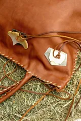 Cartera de cuero color camel, de silueta amplia y blanda, con manijas largas y terminación artesanal en los bordes. Cuenta con apliques decorativos frontales de alpaca y nácar, tientos largos colgantes y bolsillo interno.