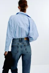 Jeans de mujer, tiro alto, corte Mom Fit con pierna slim y largo tobillero. Denim azul medio con efecto desgastado y lavado. Presenta cinco bolsillos y cierre frontal con cremallera y botón.