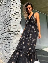 Vestido largo negro con escote en V, breteles finos y estampado floral dorado. Presenta detalles de crochet en el escote y la cintura, y un lazo con borlas en la cintura.