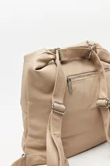 Cartera convertible en mochila, confeccionada en cuero sintético texturizado color marrón. Presenta un diseño con recortes, bolsillo frontal con cierre diagonal, herrajes metálicos plateados, asa superior y correas regulables.