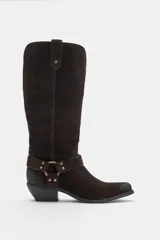 Bota alta de serraje marrón oscuro, de estilo texano, con tacón en bloque y puntera ligeramente puntiaguda. Cuenta con detalle de tiras cruzadas con tachas metálicas y una arandela en el tobillo, además de trabillas laterales en la parte superior de la caña.
