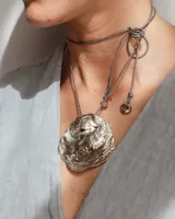 Collar artesanal con dije central de metal con textura orgánica irregular que evoca una ostra. Presenta un sistema de sujeción mediante hilos de algodón ajustables, permitiendo diversas formas de uso como gargantilla o collar largo.