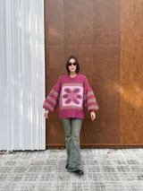 Buzo de tejido de punto con textura peluda, de corte holgado y mangas amplias. Presenta un diseño geométrico con una flor grande estampada en el centro, combinando tonos fucsia, rosa y marrón en un patrón de bloques y rayas.