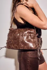 Cartera bandolera de cuero sintético marrón con efecto desgastado, detalles de cordones cruzados por ojalillos metálicos en los laterales y placa metálica con el logo de la marca.