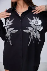 Camisa negra de manga larga con cuello clásico y botones frontales. Presenta dos grandes apliques bordados con lentejuelas y cuentas en forma de flores y hojas en la parte delantera.