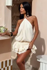 Vestido corto color beige, con escote halter, espalda descubierta y falda con volados en capas.
