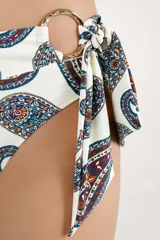 Calzón de bikini tipo faja de tiro alto, color blanco con estampado paisley en tonos azul, rojo y mostaza. Cuenta con una argolla metálica decorativa y un lazo lateral.