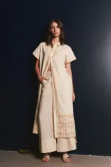 Vestido camisero de lino color lila con bordados rojos en contraste. Presenta un diseño de largo midi con cuello camisero, cierre frontal de botones y mangas cortas. Los detalles bordados recorren la botonadura, los puños y el ruedo de la prenda.