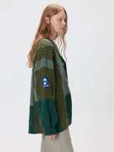 Cardigan oversize de tejido de punto grueso con diseño de bloques de color en tonos tierra. Presenta cuello en V, cierre frontal con botones, mangas largas con terminación abullonada y parches decorativos en el pecho y hombro.