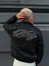 Campera de cuero sintético con textura rugosa, diseño tipo bomber con puños y cintura elásticos. Presenta un bordado grande en la espalda con tipografía cursiva en color blanco.
