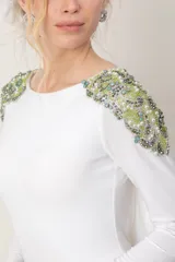 Vestido largo blanco de manga larga, con escote redondo al frente, espalda baja descubierta y detalles de bordado de piedras y pedrería en tonos verdes sobre los hombros.