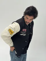 Campera estilo varsity con cuerpo negro y mangas blancas. Presenta parches bordados en el pecho y las mangas, cierre frontal con botones a presión y puños, cuello y ruedo acanalados con rayas blancas y negras.