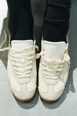 Zapatilla deportiva estilo blucher de color blanco, con detalles en gris claro, una franja lateral blanca y una talonera azul. Cuenta con cordones, punta redonda y suela de goma con tachuelas.