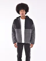 Campera con capucha, diseño de bloques de color. La parte superior (hombros y capucha) es de tela lisa negra, mientras que el cuerpo y las mangas son de tejido polar o sherpa gris oscuro. Cierre frontal.