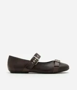 Zapatilla tipo bailarina color marrón oscuro, confeccionada en poliuretano con acabado mate. Presenta punta redondeada, suela plana y dos tiras ajustables con hebillas metálicas plateadas y ojales decorativos sobre el empeine.