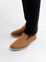 Zapatilla tipo slip-on color marrón, con diseño minimalista, elásticos laterales para mayor comodidad y suela blanca con detalle de costura visible.