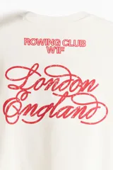 Remera boxy de algodón color blanco, con cuello redondo acanalado, mangas cortas y hombros caídos. Presenta un pequeño estampado rojo en el pecho con las inscripciones 'ROWING CLUB W1F'.