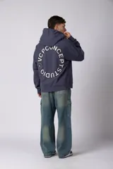 Buzo canguro oversize de frisa de algodón premium en color azul grisáceo. Presenta un diseño con recortes frontales, bolsillo tipo canguro y un pequeño logo circular estampado en el pecho. Cuenta con puños y ruedo acanalados.