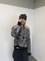 Buzo con cierre frontal completo, confeccionado en tejido de punto grueso de algodón. Presenta un diseño estampado integral con motivos gráficos y tipografía estilo gótico en tonos gris y negro, capucha integrada, puños y ruedo acanalados en color negro sólido.