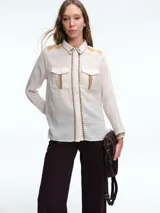 Camisa de color crudo con detalles de bordados en tonos mostaza y marrón en el cuello, hombros, bolsillos frontales y puños. Presenta cierre frontal con botones y dos bolsillos tipo cargo en el pecho.